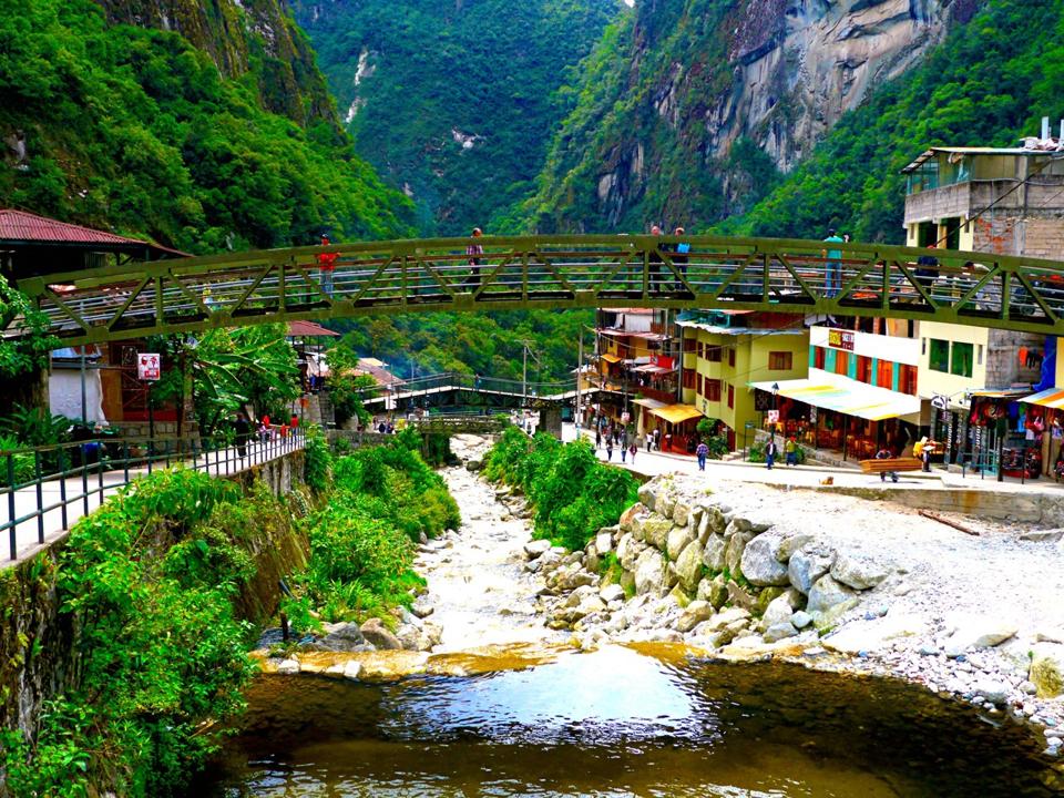 A pedestrian bridge over the river in Aguas Calientes; there are a few buildings on either side and steep green mountains in the background.