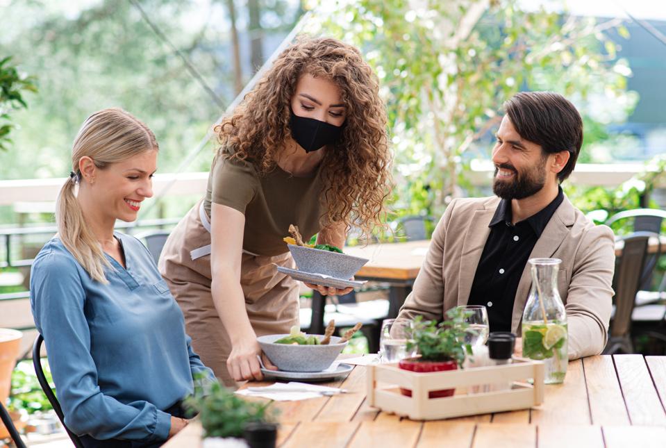 Waitress with face mask serving happy couple outdoors on terrace restaurant.