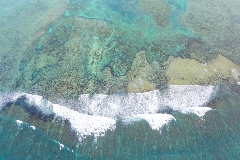 Shot from above, waves break on a coral reef beyond which is calm water