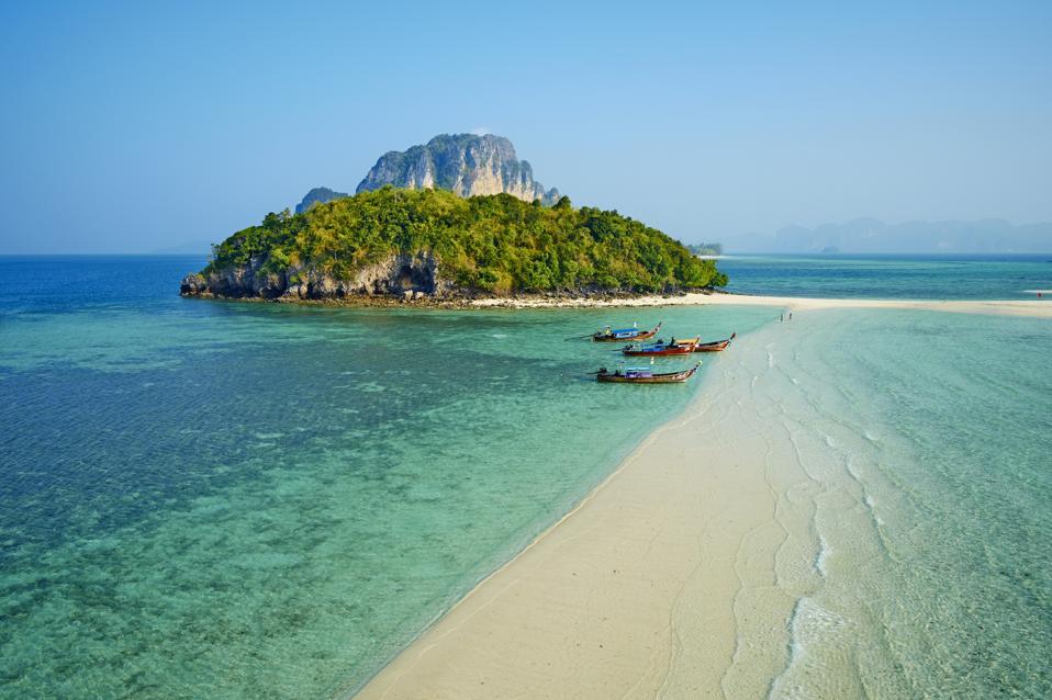Four longtail boats are tied up beside a long spit of sand that is barely underwater. In the background is a green-treed island with a tall rocky center.