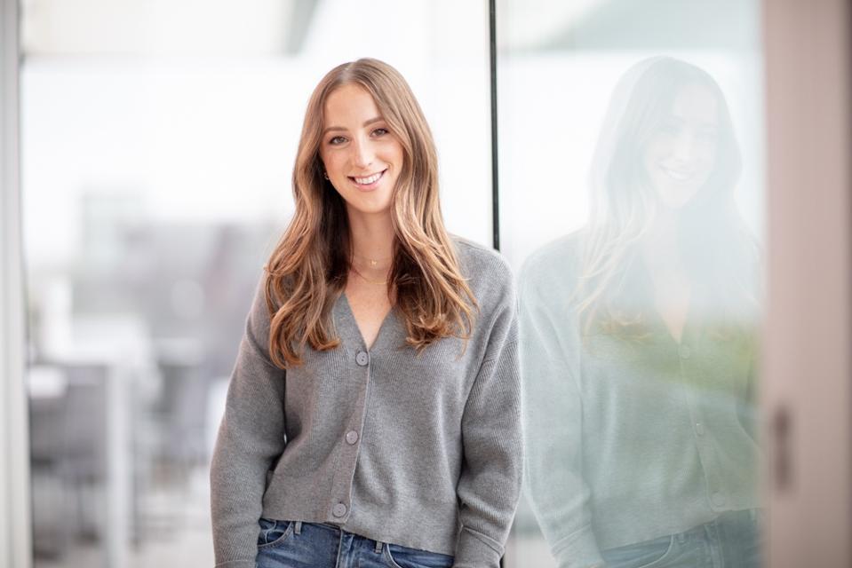 a brunette woman wearing grey cardigan standing in front of floor to ceiling window