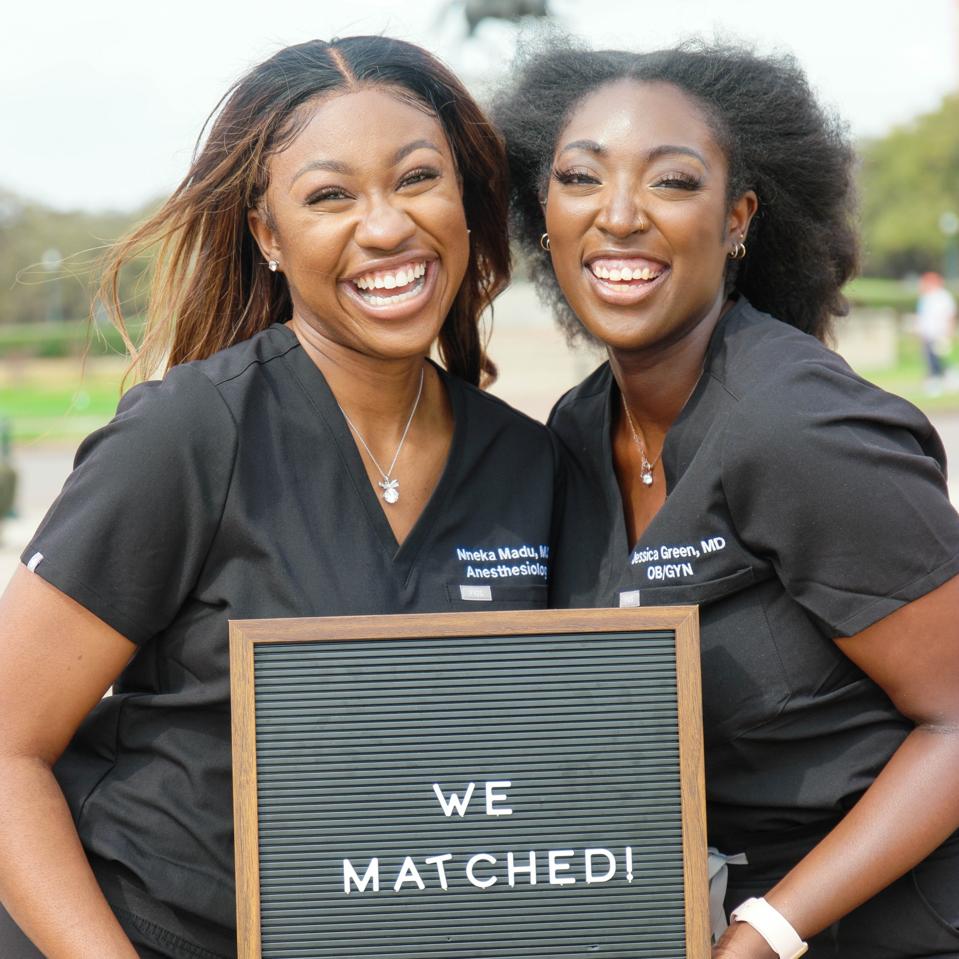 Nneka Madu and Jessica Green holding a match sign and smiling in their medical wear.