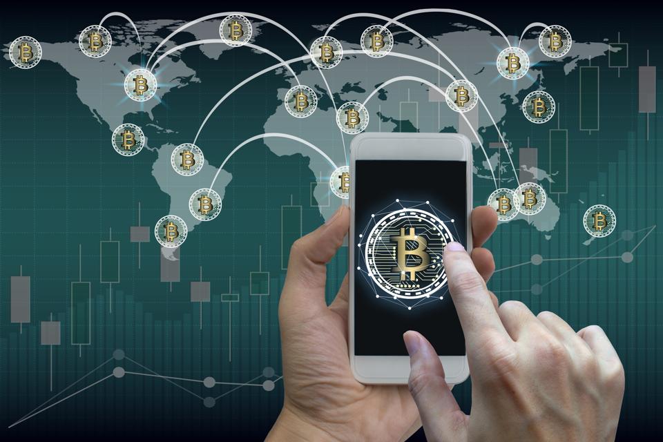 Close-Up Of Hand Using Mobile Phone With Bitcoin Symbol Against World Map