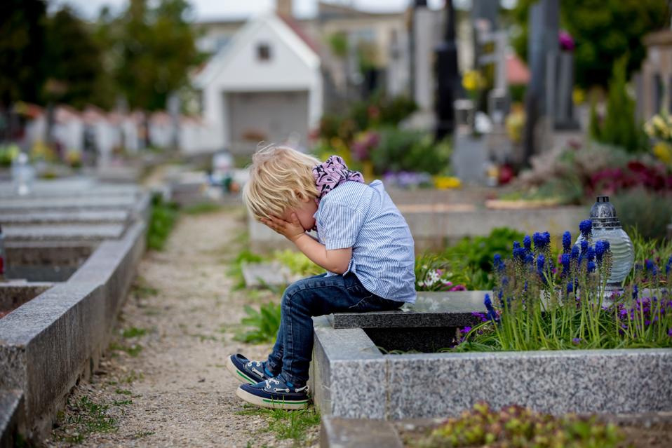 Little toddler boy, sitting on a grave in cemetery, sad and lonely, springtime