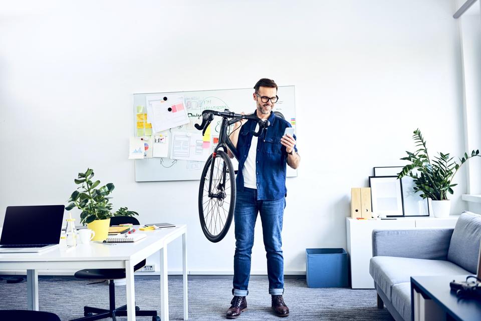 Smiling businessman carrying bicycle in office looking at smartphone