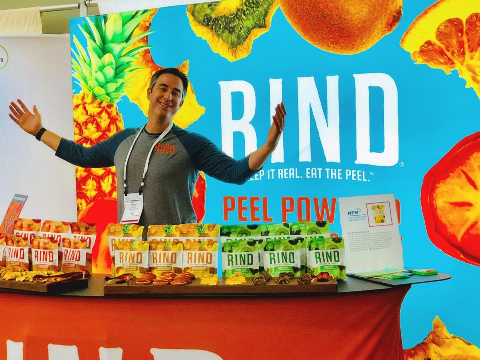 Weiss stands in front of a RIND banner with a table full of RIND snacks.