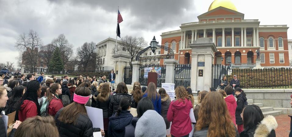 A person addresses a crowd of young people in front of the Massachusetts State House.