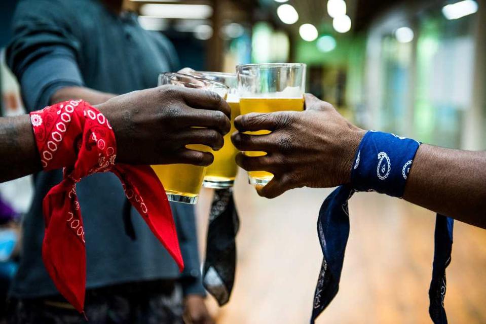 Three Black men toast with beers while wearing bandanas around their arms with colors that show their rival gang affiliations