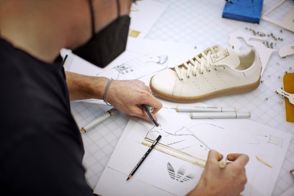 A man draws shoe designs next to the white Mylo Adidas Stan Smith shoe.