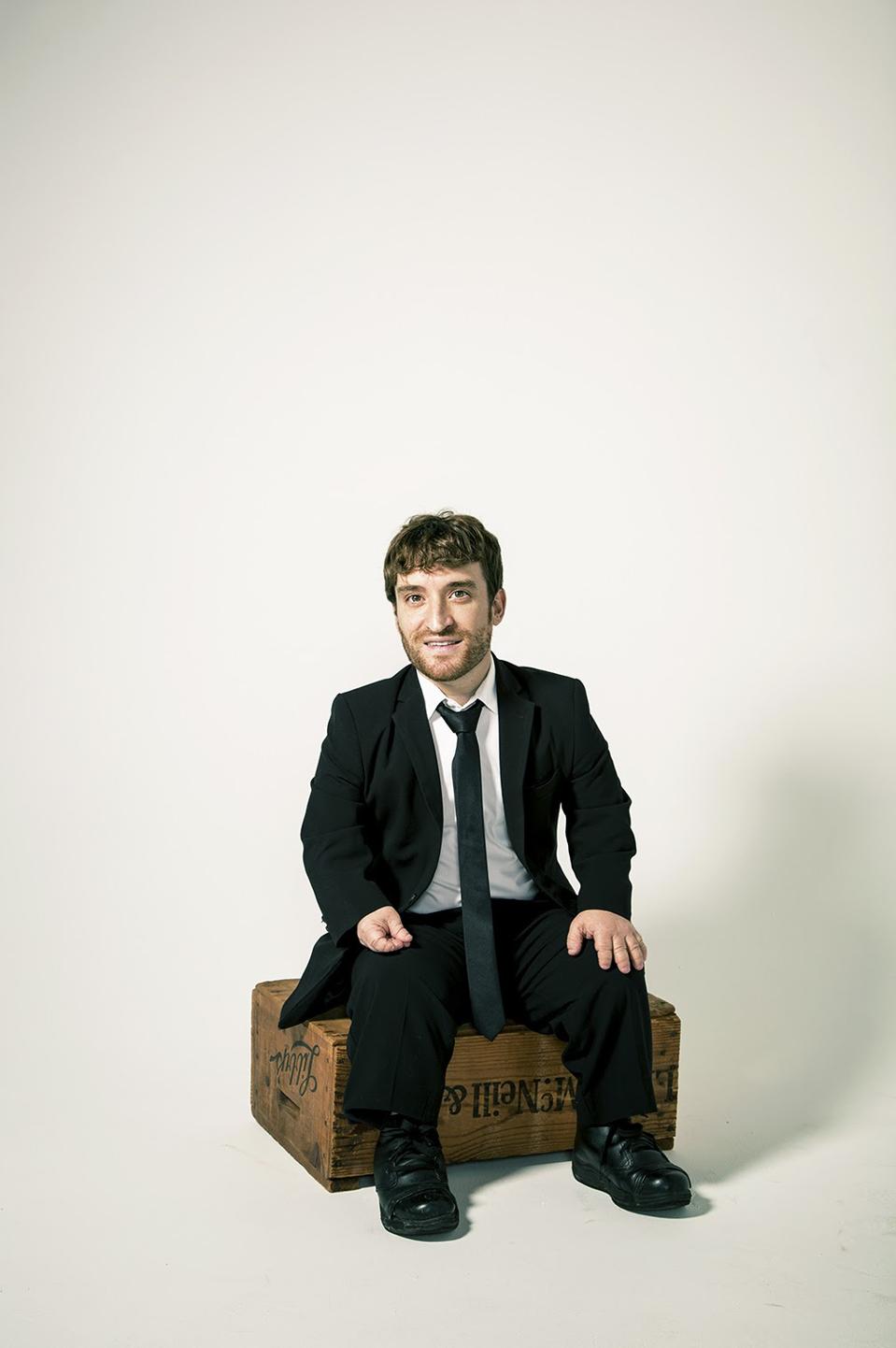 A white man, who is a little person, sits on a wood box wearing a suit. 