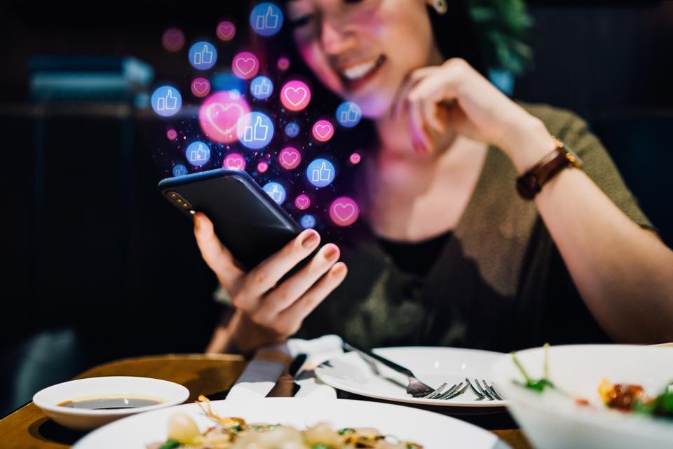 Smiling young Asian woman using smartphone on social media network application while having meal in the restaurant, viewing or giving likes, love, comment, friends and pages. Social media addiction concept