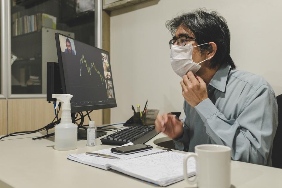 Worker alone in office adjusts his mask during a video conference with remote colleagues