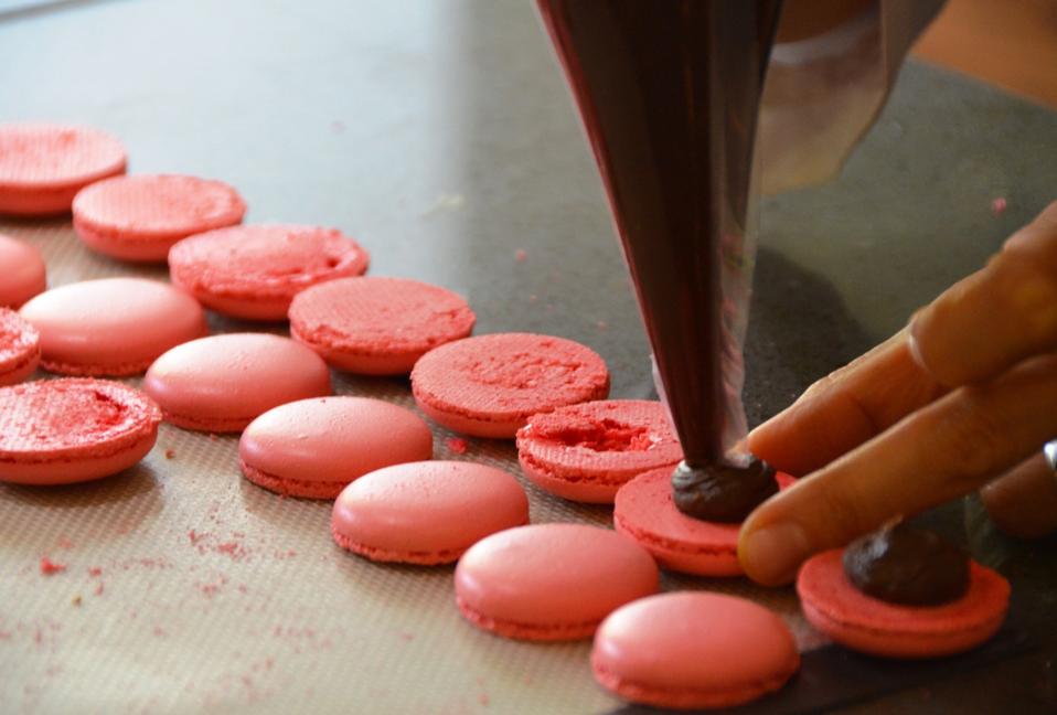 Piping Dark Chocolate Ganache on the shells of the cooled soft pink macarons is shown in this photograph from La Cuisine Paris. 