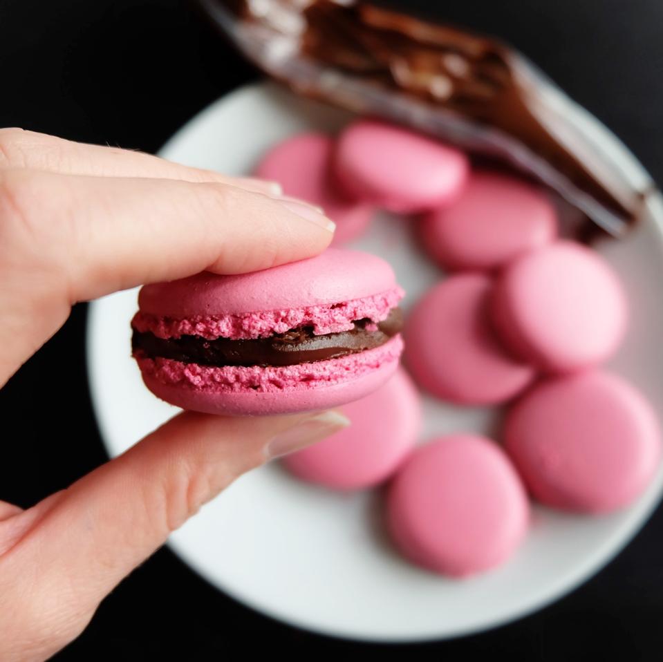 A woman is holding a soft pink macaron filled with dark chocolate ganache in this photo from La Cuisine Paris.