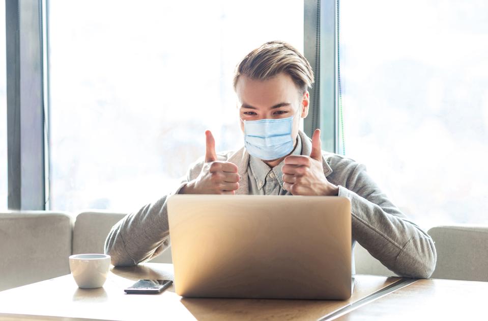 Working at home safe in quarantine time. Young satisfied man with surgical medical mask, sitting, looking at laptop screen with thumbs up.