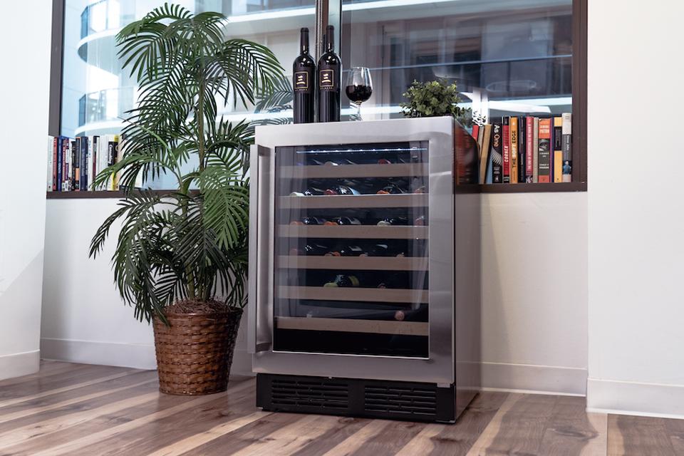 Wine cooler in a room with a plant next to it and a glass of wine on top. In the background are book and a window