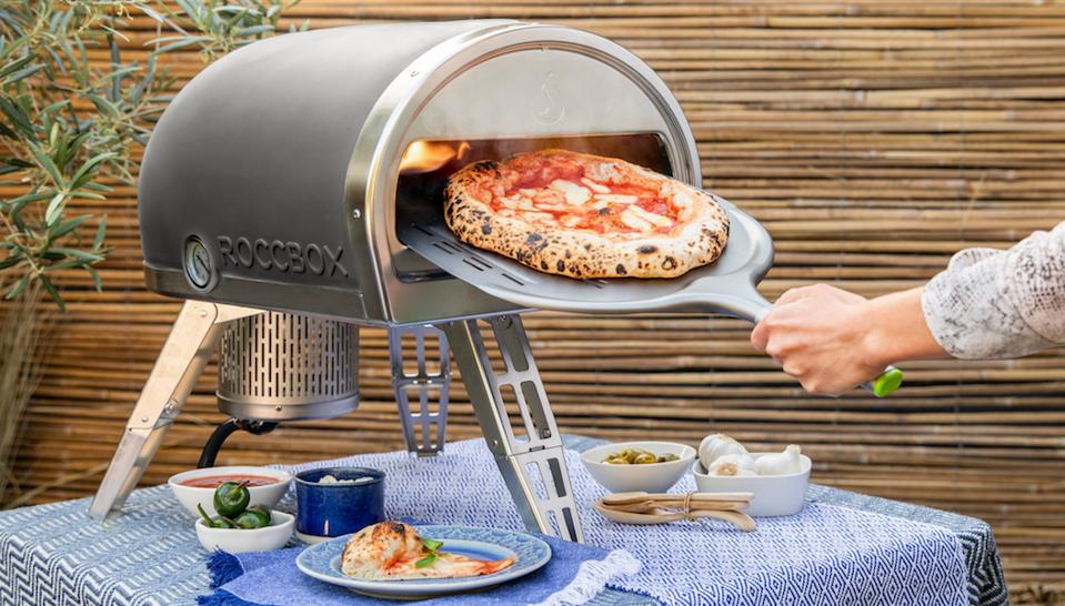 Gozney Roccbox Portable Pizza Oven on a table outside with a hand holding a pizza tray