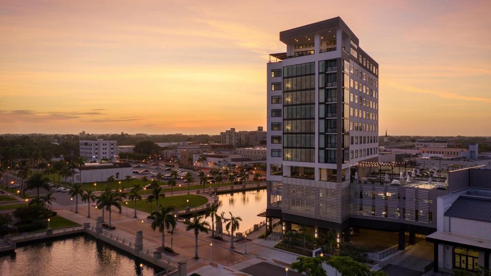 A tall hotel lined with windows standing above a river, the sun is setting