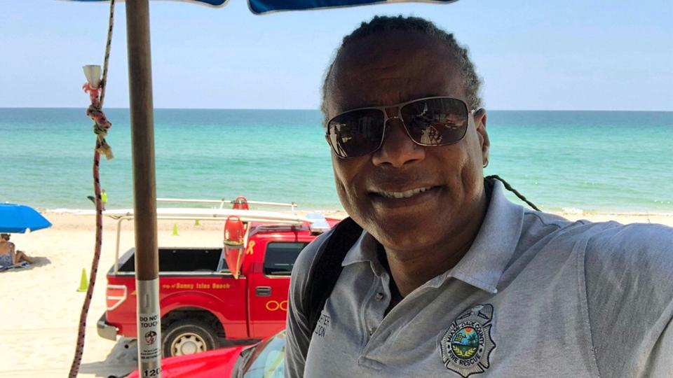 A Black Firefighter standing in front of his fire truck on the beach in Miami, Florida.