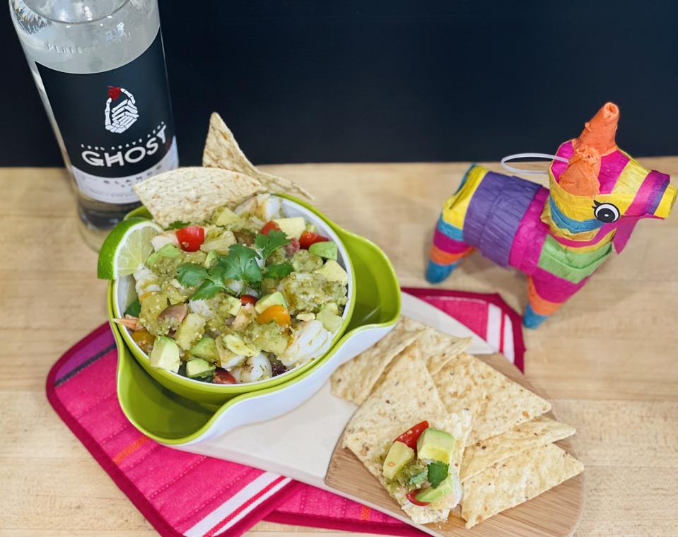 A bowl of grilled shrimp ceviche is shown in a bowl with a garnish of fresh lime, cilantro and tortilla chips. In the background is a colorful miniature piñata and a bottle of Ghost Tequila.