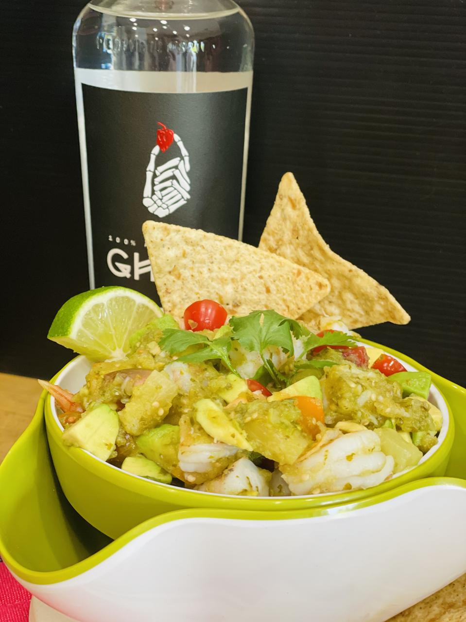 A green and white bowl of grilled shrimp ceviche made with tomatillo salsa, avocado, grape tomatoes, pineapple tidbits and spicy Ghost Tequila is shown in front of a bottle of Ghost Tequila.