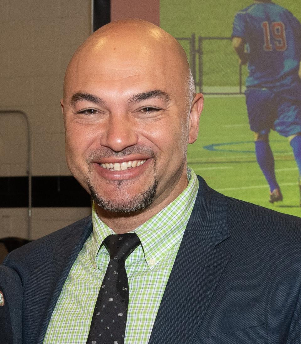 Bald headed man smiling into camera while wearing suit and tie, seen from chest up.