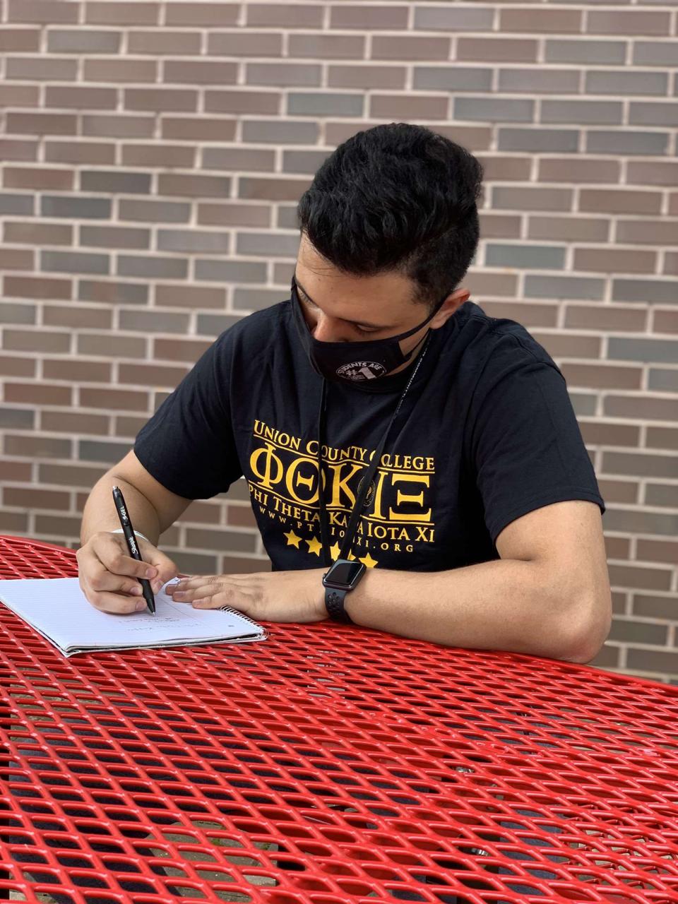 Masked UCC student in a t-shirt writing with a pen and notebook.