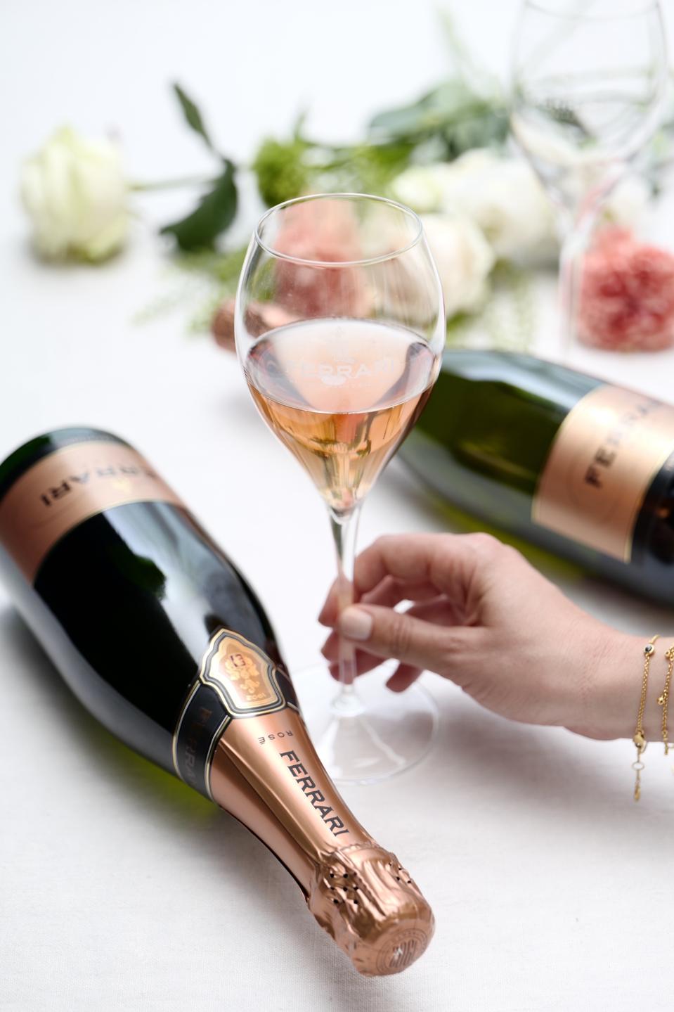 A woman holds a glass of Ferrari Trento sparkling rosé in between two bottles.