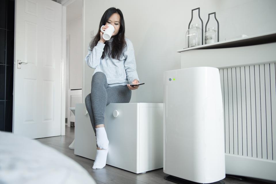 A young Asian woman controls the power of her smart air unit from her mobile phone as she sits on a storage bench and enjoys a cup of tea in the bedroom
