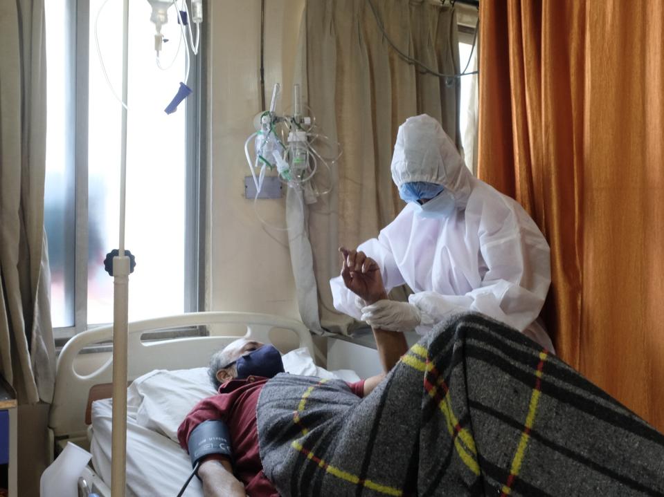 A doctor dressed in head to toe PPE stands bedside a patient in a hospital. 