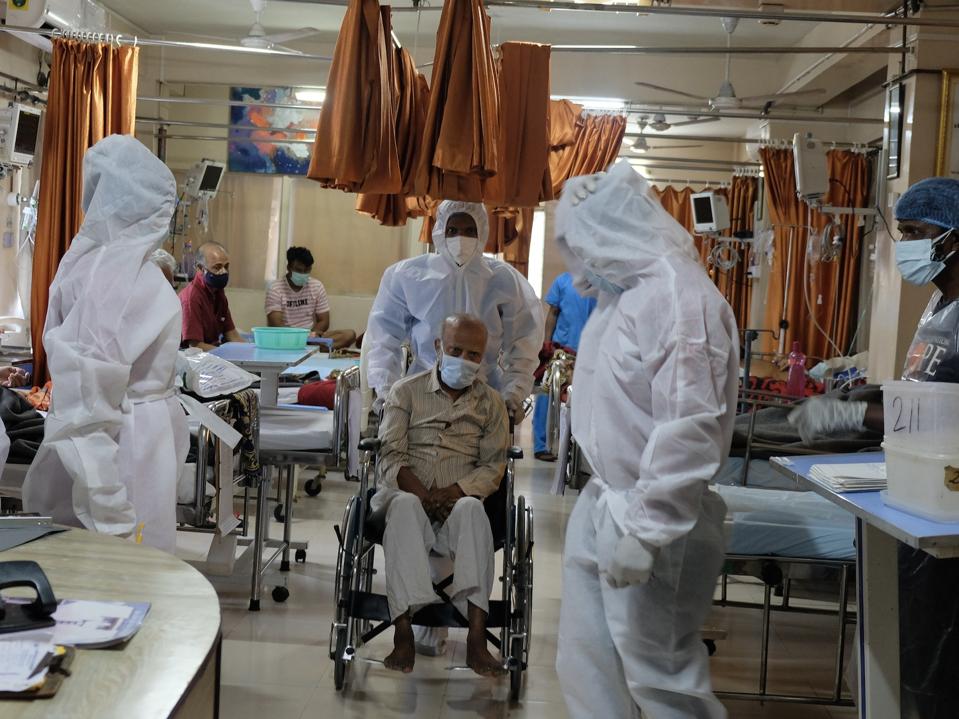 A man with a mask sits in a wheelchair in a hospital filled with staff in PPE.