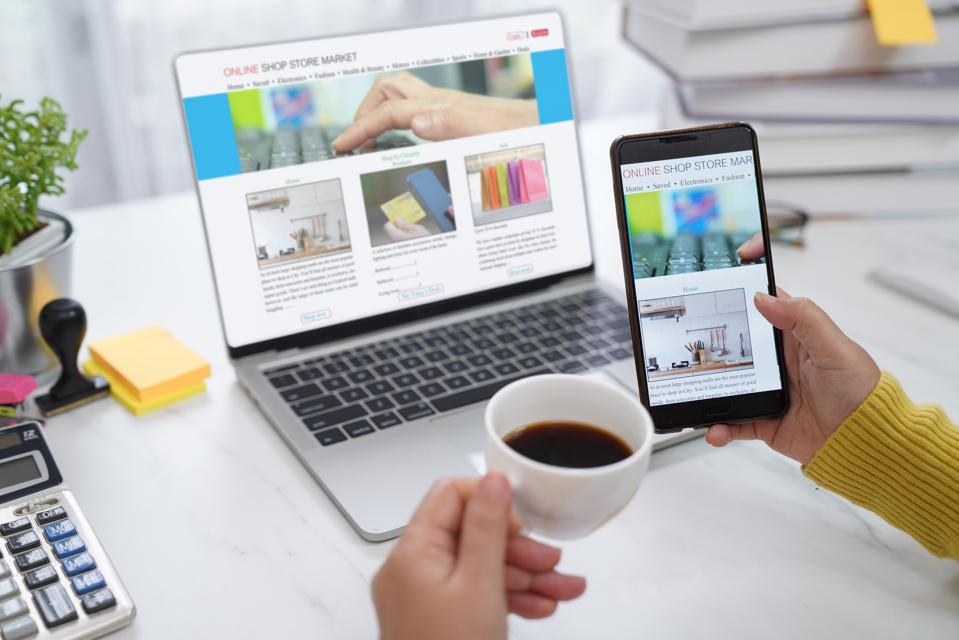 Hands holding a phone a phone and a coffee cup with a laptop screen open in front showing an online shop