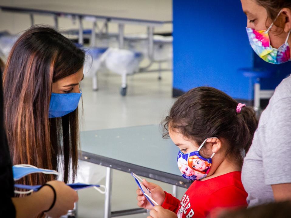 Sharma bends down to talk w a little girl standing next to her mother. All wear face masks