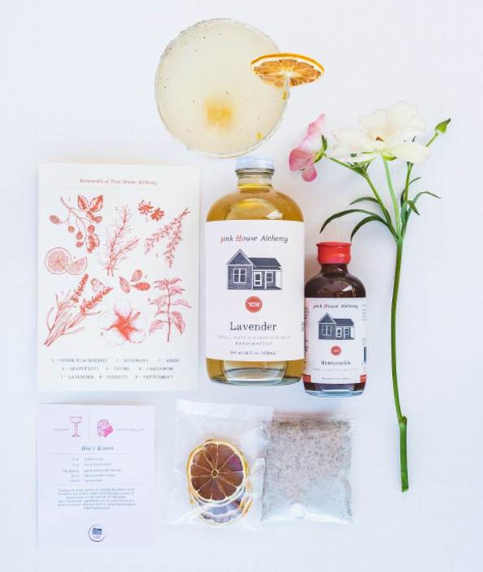 This is a photo of the contents of the Pink House Alchemy Bee's Knees cocktail kit which will be available through their website through August. The kit contains a recipe card, Lavender simple syrup, Honeysuckle bitters, dehydrated lemon slices, and Lavender Bee Pollen Rimming Sugar.