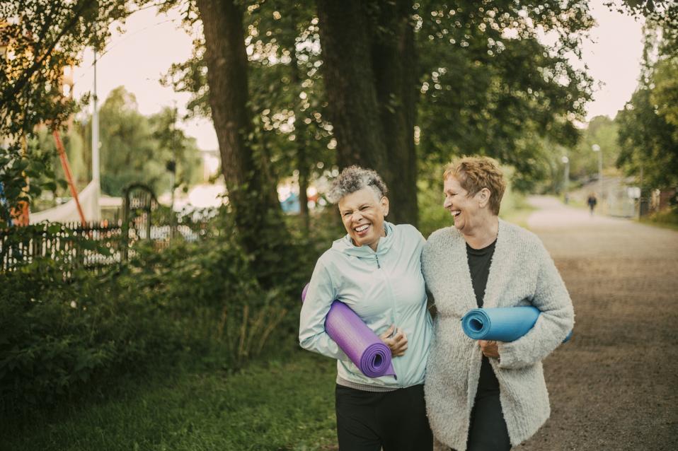 Smiling female friends holding exercise mat while walking on footpath