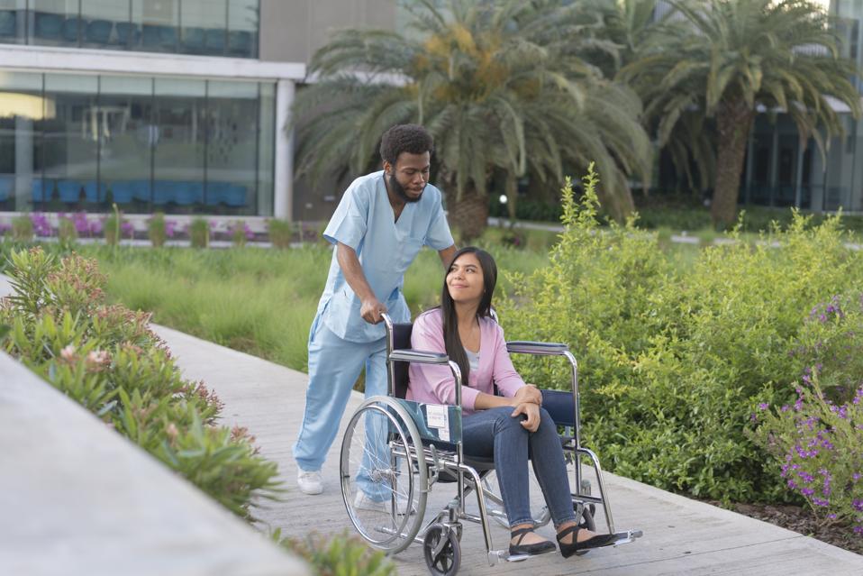 Black male nurse assists a young woman patient through a hospital garden.
