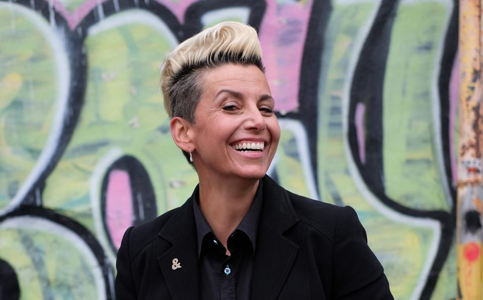 MJ DePalma with blonde short hair, styled in a pompadour with forearm tattoos showing while wearing black blazer and smiling standing in front of graffiti in Los Angeles 2021