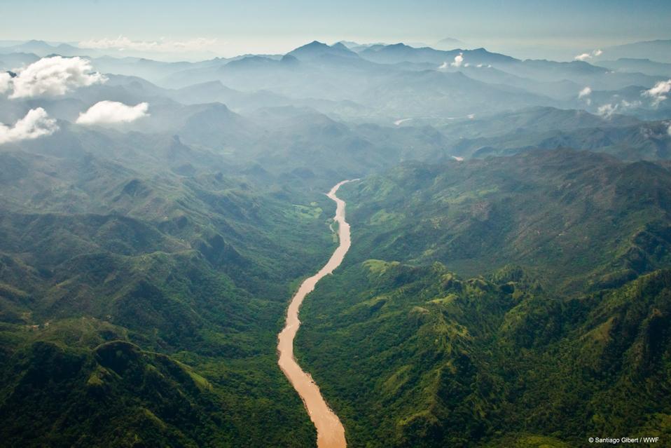 An aerial view of a thin brown river winding through jungle-covered hills