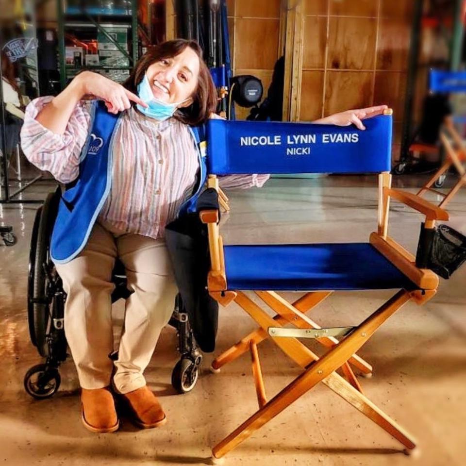 A white woman in a wheelchair poses next to a chair that says ″Nicole Lynn Evans.″