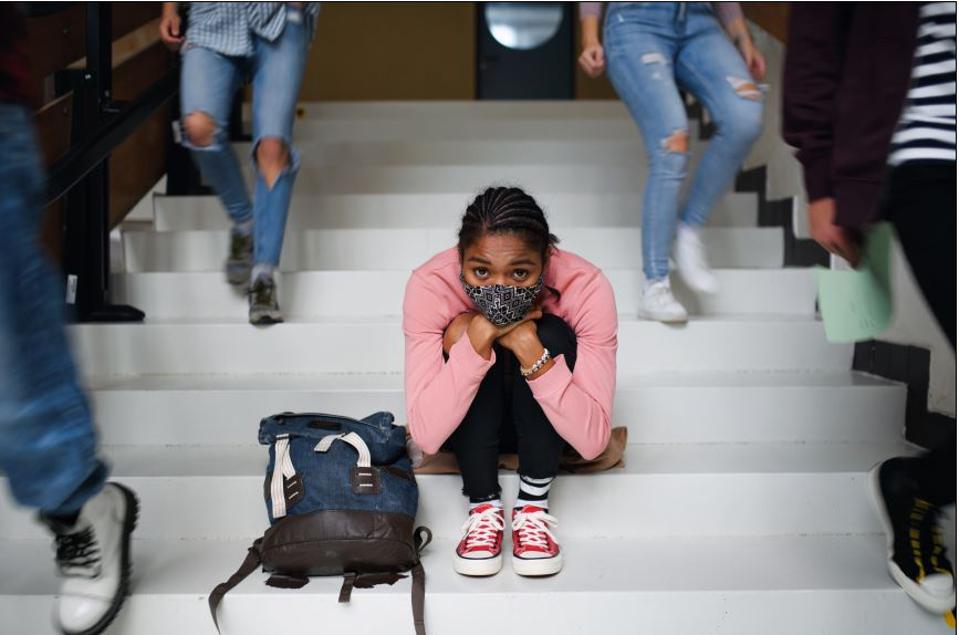 Girl wearing a mask, huddled up, sitting on stairs while people walk around her.