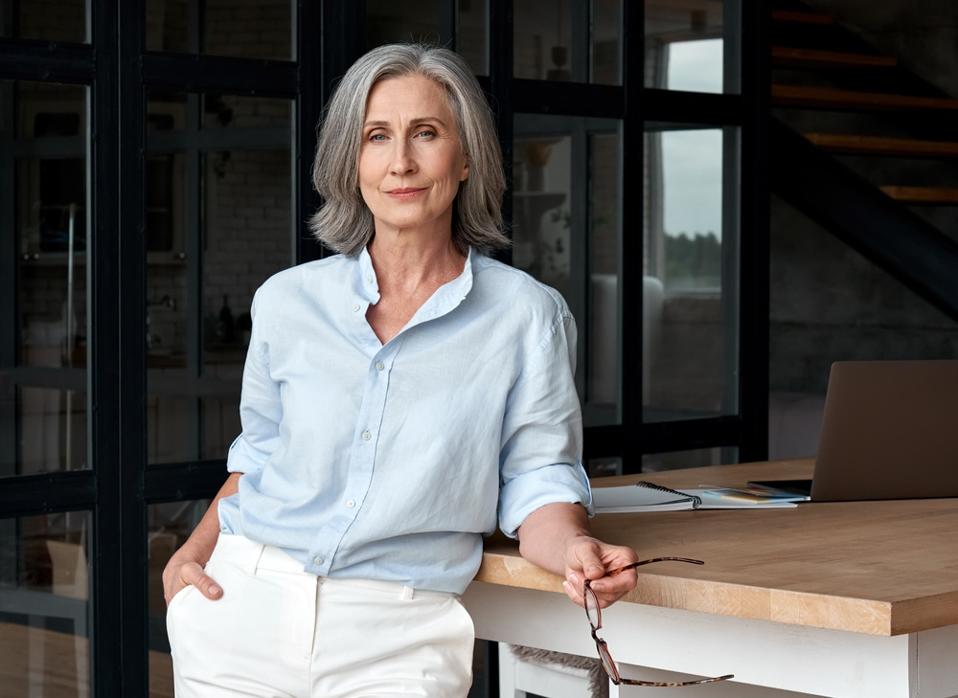 Older female leader in modern glass office gazes confidently to camera.