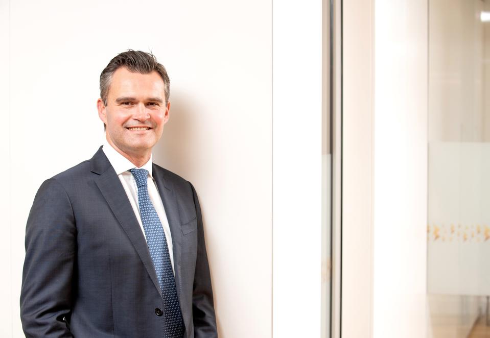 Tony Sacre, white middle-aged man in suit and tie, brushed back hair, smiling at camera