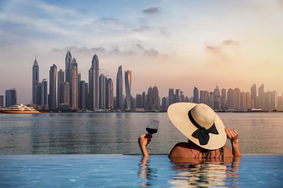 A woman with a hat stands at the edge of a pool and enjoys the view to the skyline of the Dubai Marina