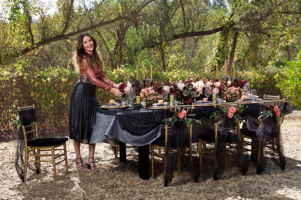 A brunette stands next to a stable that has candles, placesettings, flowers and a black tablecloth