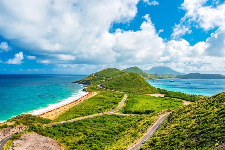 Green hills and sandy beaches of St. Kitts surrounded by various shades of blue ocean, with the hills of the island of Nevis in the background.