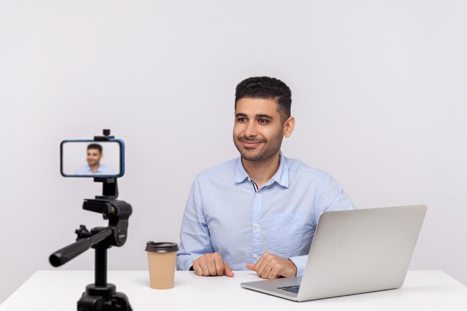 Businessman creating video in workplace, smiling at phone on tripod.
