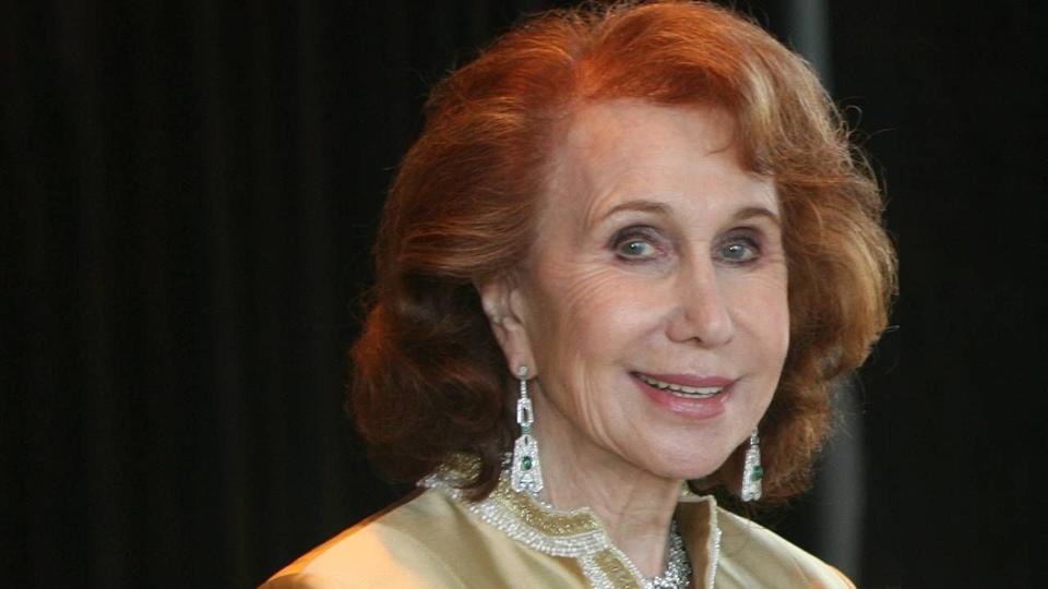 An older woman with red hair wearing diamond earrings and a gold jacket. 