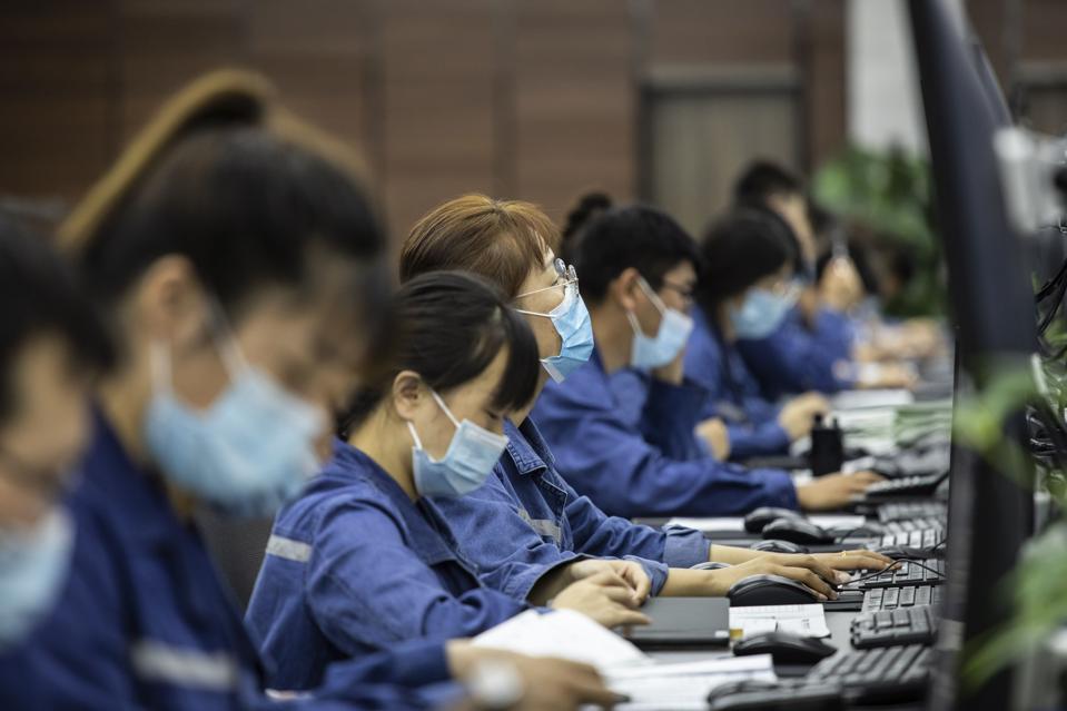 Nearly a dozen Chinese workers in masks and blue work clothes sit behind computer screens.