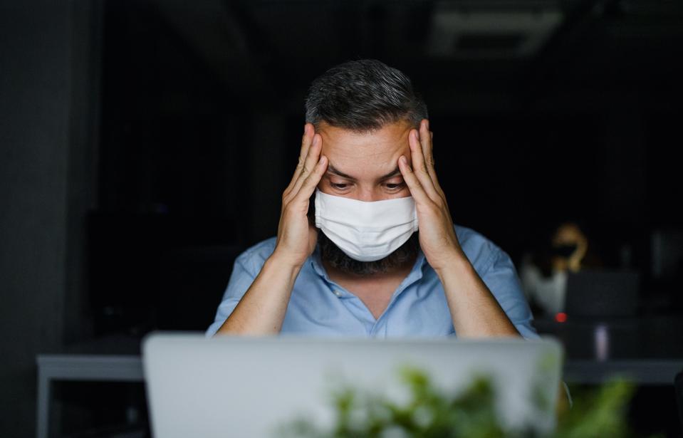 Frustrated mature man with face mask sitting indoors in office, working.