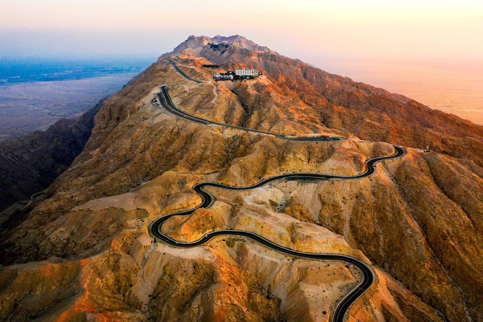 A black paved road winds up a rocky, golden mountain with a whilte building near the summit.